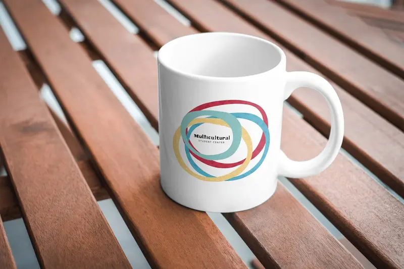 Merchandise for the Multicultural Student Center: a white mug with the four-circle graphic on a wooden table.