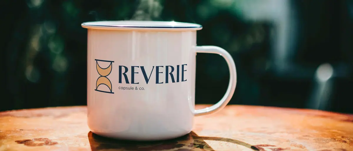 Enamel mug for Reverie Capsule & Co.: the hourglass logo on a white camp mug, steam rising, on a sunlit table.