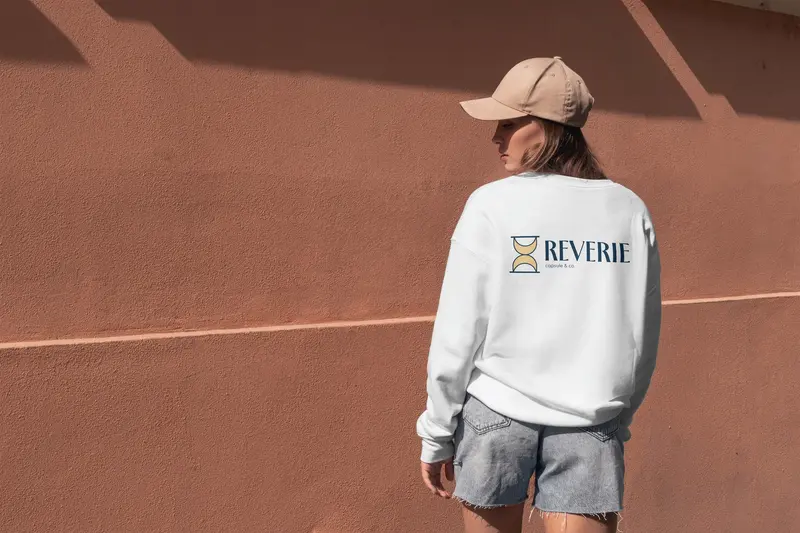 Apparel for Reverie Capsule & Co.: a white sweatshirt with the logo, worn against a terracotta wall.